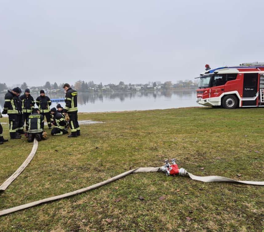 Feuerwehrleute üben am See mit einem Feuerwehrwagen, Schläuchen und einem Hydranten auf dem Gras.