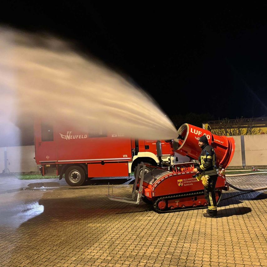 Ein Feuerwehrmann benutzt eine rote Schaumkanone, um nachts Wasser zu sprühen, mit einem roten Feuerwehrwagen im Hintergrund.