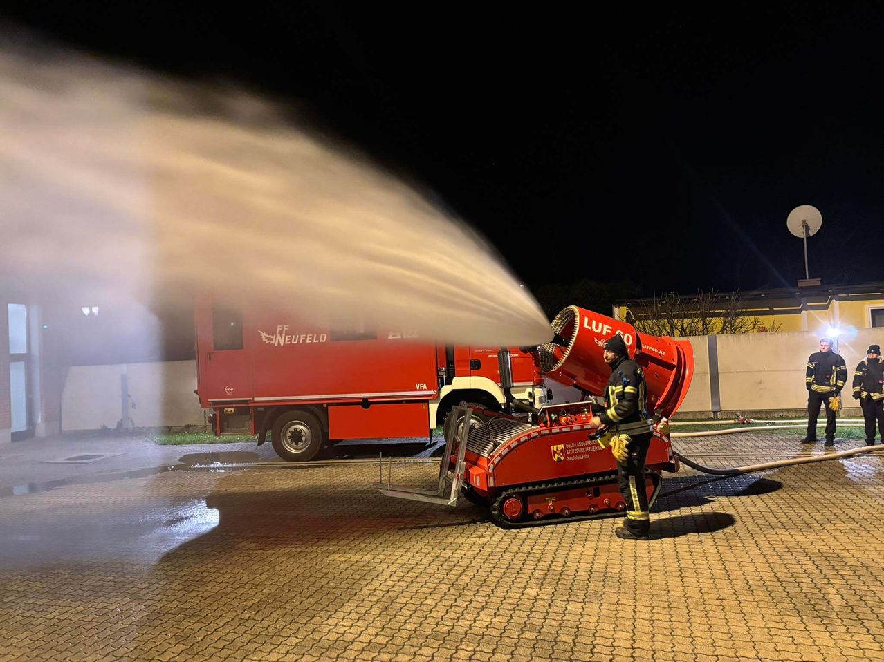 Ein Feuerwehrmann benutzt eine rote Schaumkanone, um nachts Wasser zu sprühen, mit einem roten Feuerwehrwagen im Hintergrund.