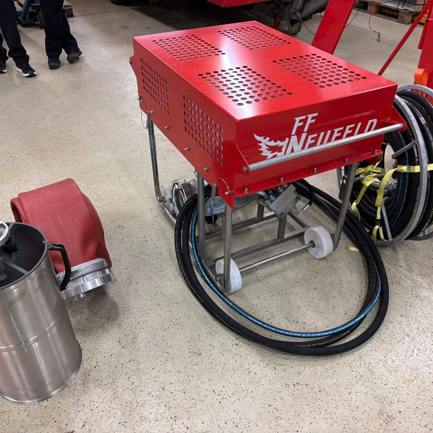 Eine rote FF NEUFELD-Maschine mit einem roten Griff und Schläuchen auf einem sauberen Boden. Ein Edelstahlbehälter befindet sich links.