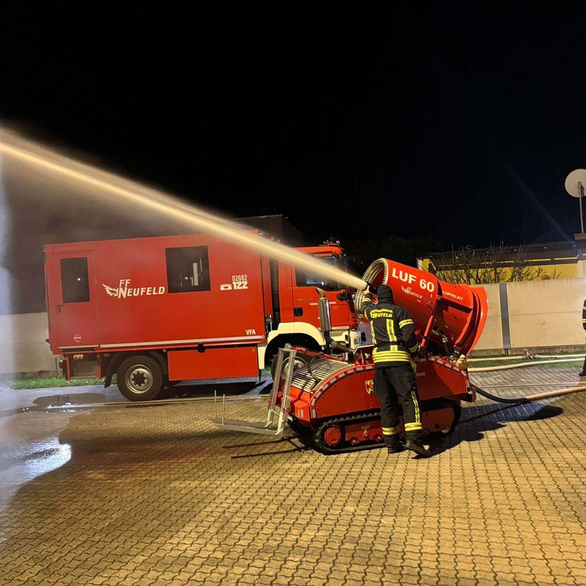 Ein Feuerwehrmann bedient einen Feuerwehrwagen, der nachts Wasser sprüht. Der LKW ist mit 'FF NEUFELD' beschriftet, und der Feuerwehrwagen trägt die Bezeichnung 'LUF 60'.