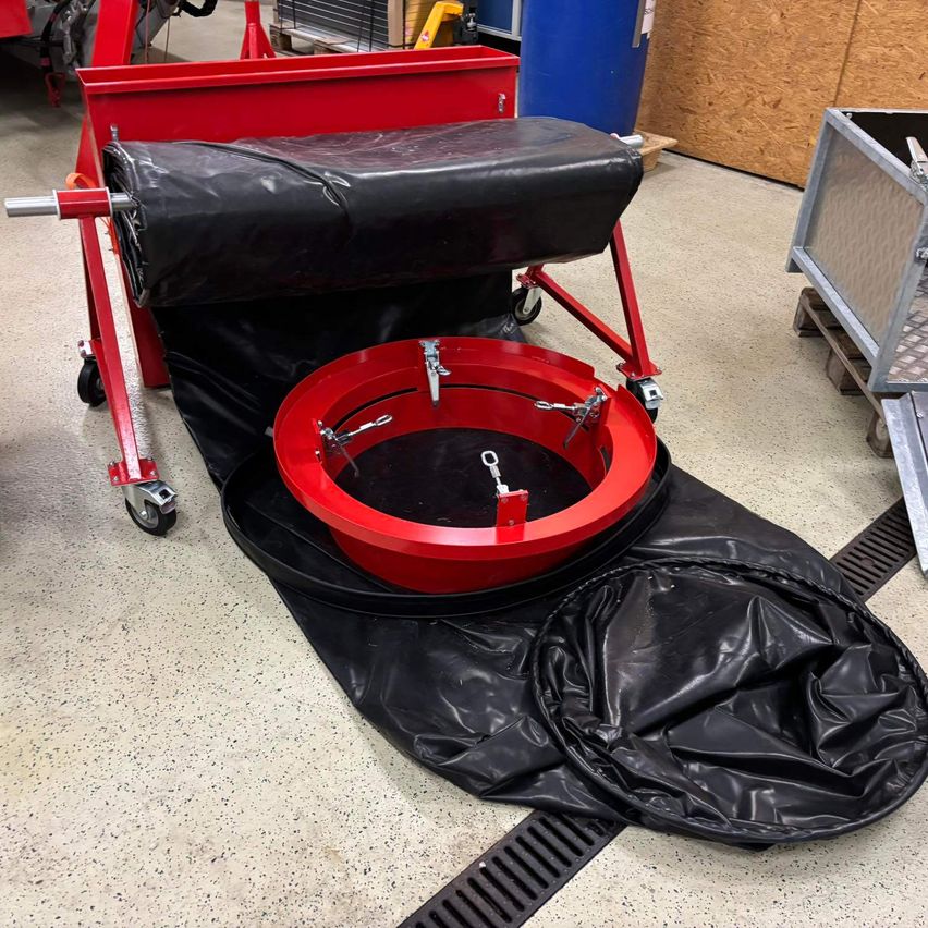 Ein rotes kreisförmiges Objekt mit Metallbefestigungen ruht auf einer schwarzen Plane, positioniert auf einer Plattform mit Rädern. Die Plattform ist mit schwarzem Material bedeckt, und im Hintergrund sind ein Holzpalette, ein blauer Fass und andere industrielle Objekte zu sehen.