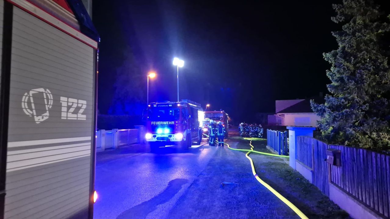 Feuerwehrleute sind nachts bei einem Vorfall, mit eingeschalteten Fahrzeuglichtern. Sie sind in der Nähe eines Gebäudes mit einem gelben Schlauch.