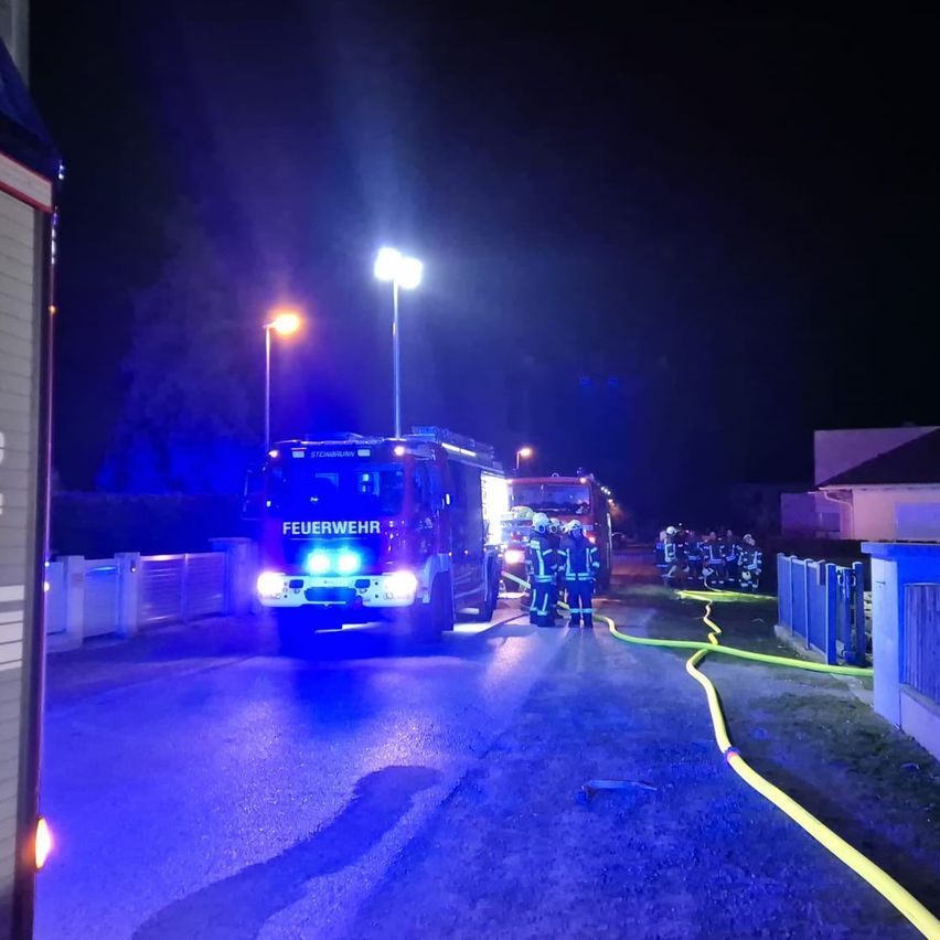 Feuerwehrleute sind nachts bei einem Vorfall, mit eingeschalteten Fahrzeuglichtern. Sie sind in der Nähe eines Gebäudes mit einem gelben Schlauch.