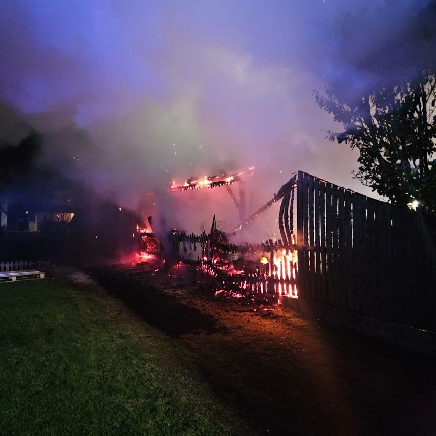 Ein Haus steht bei Nacht in Flammen, wobei Rauch in den Himmel steigt. Das Feuer breitet sich auf einen Holzzaun aus.
