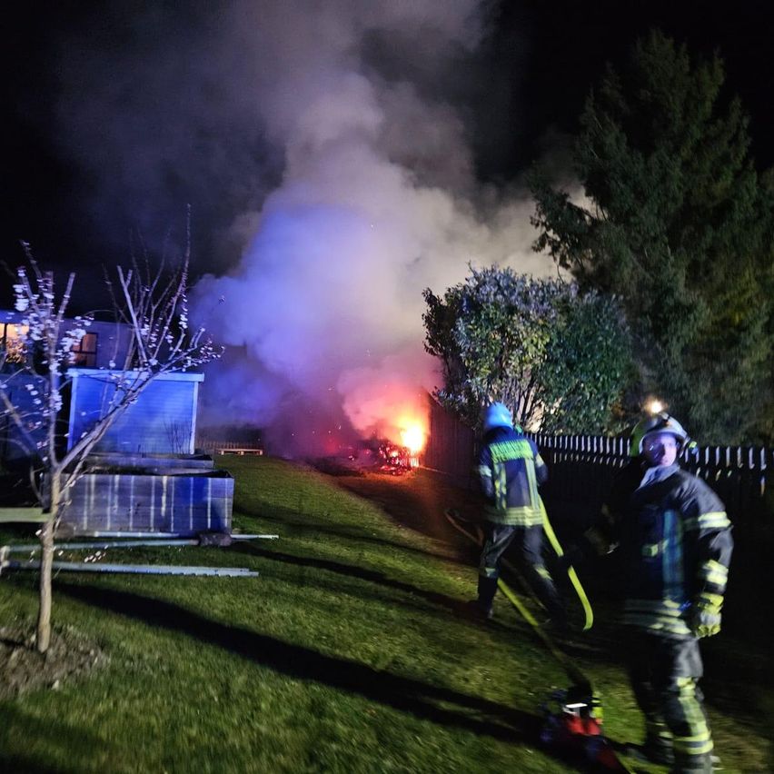 Feuerwehrleute löschen ein Feuer bei Nacht. Flammen und Rauch steigen auf und beleuchten die Szene gegen einen dunklen Himmel. Bäume und ein Zaun sind in der Nähe.