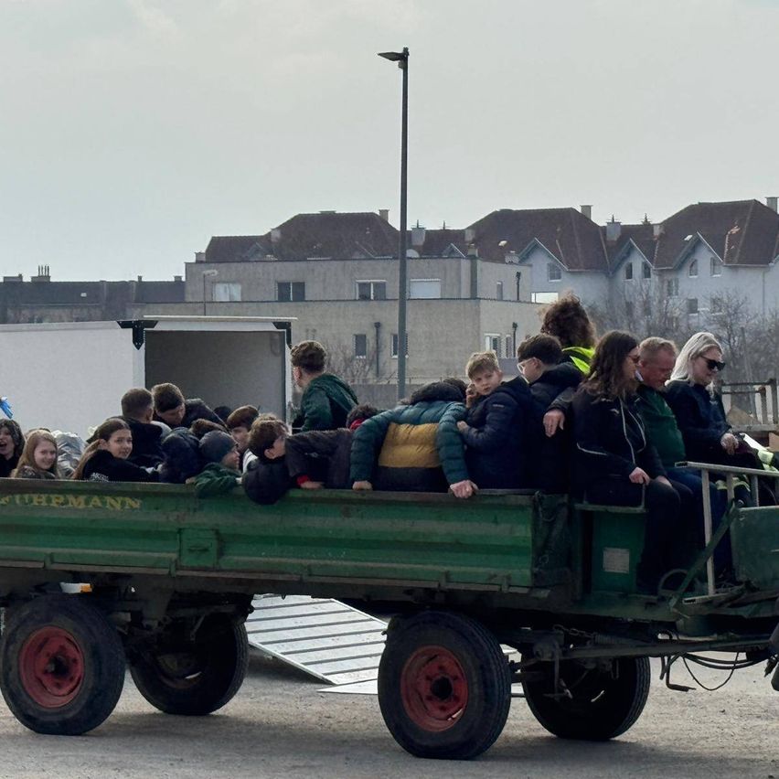 Eine Gruppe von Menschen, einschließlich Kindern, ist in einem grünen Anhänger mit einer weißen Rampe gedrängt. Sie sind von Gebäuden, einer Straßenlaterne und einem bewölkten Himmel umgeben.