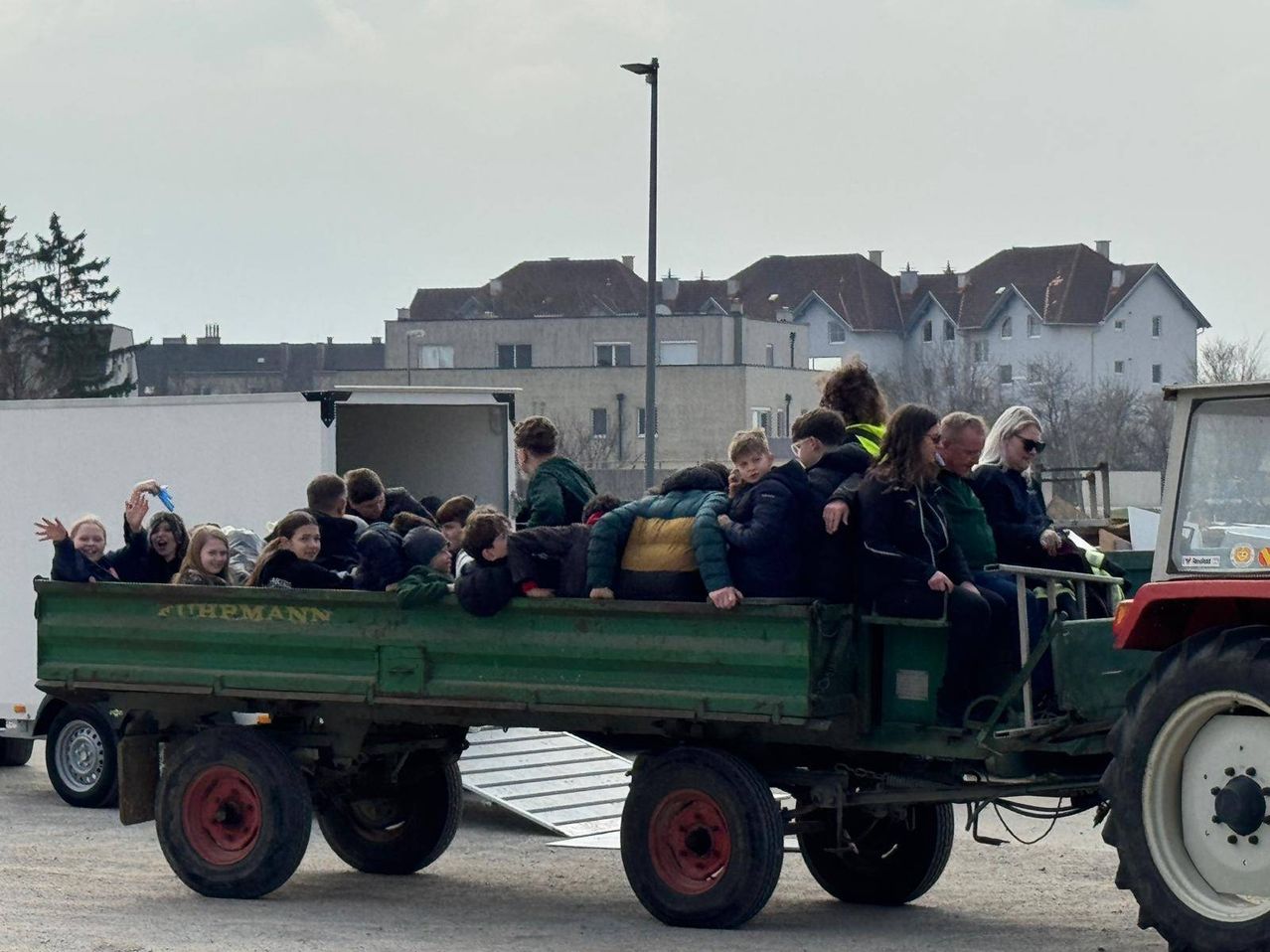 Eine Gruppe von Menschen, einschließlich Kindern, ist in einem grünen Anhänger mit einer weißen Rampe gedrängt. Sie sind von Gebäuden, einer Straßenlaterne und einem bewölkten Himmel umgeben.