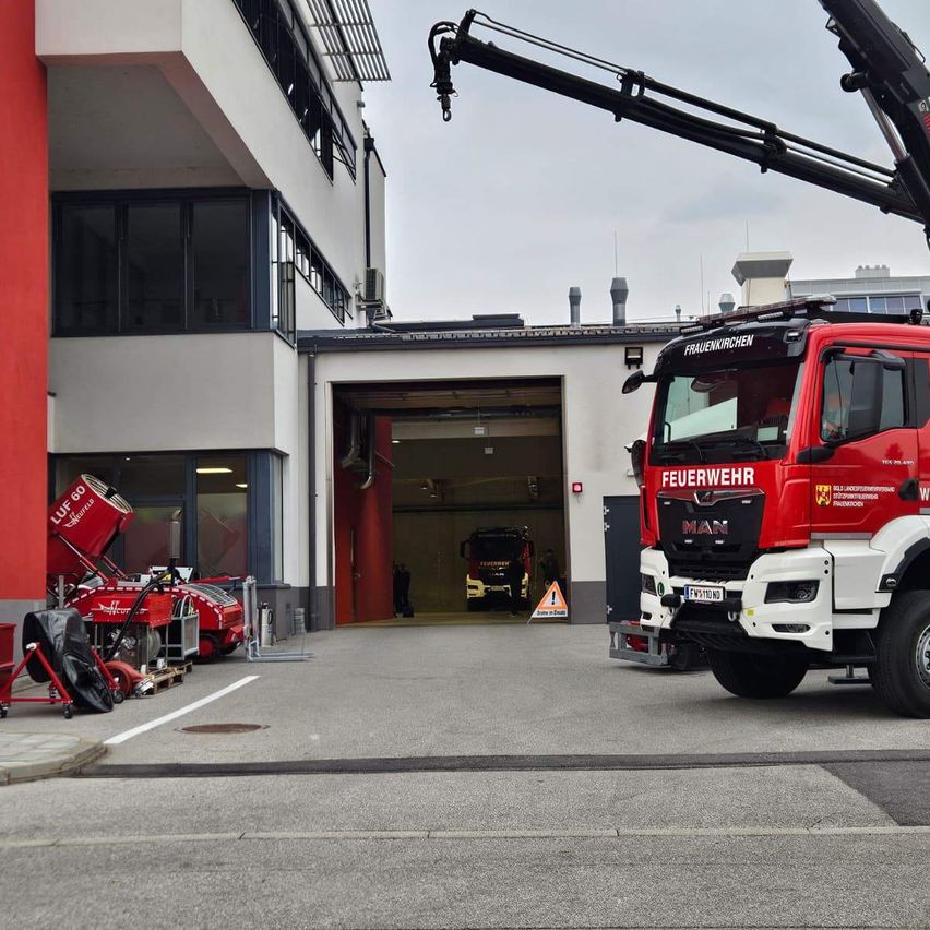 Ein roter Feuerwehrwagen steht vor einem Gebäude mit einer roten Säule und einer offenen Garagentür. Zwei weitere Feuerwehrwagen sind in der Garage geparkt.