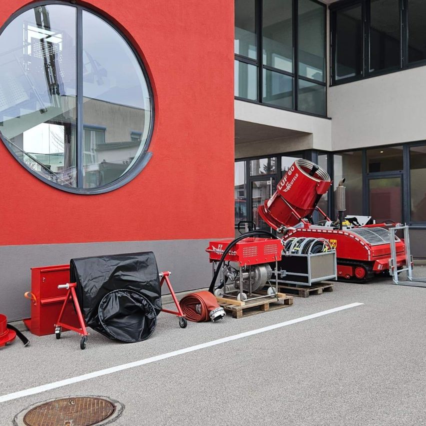 Rote Maschinen, darunter ein roter Container und ein roter Schubkarren, stehen vor einem roten Gebäude mit einem runden Fenster.