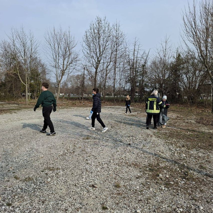 Fünf Menschen gehen auf einem Kiesweg. Einige tragen Jacken, und einer hält einen Müllsack. Bäume umgeben sie, und der Boden ist kahl.