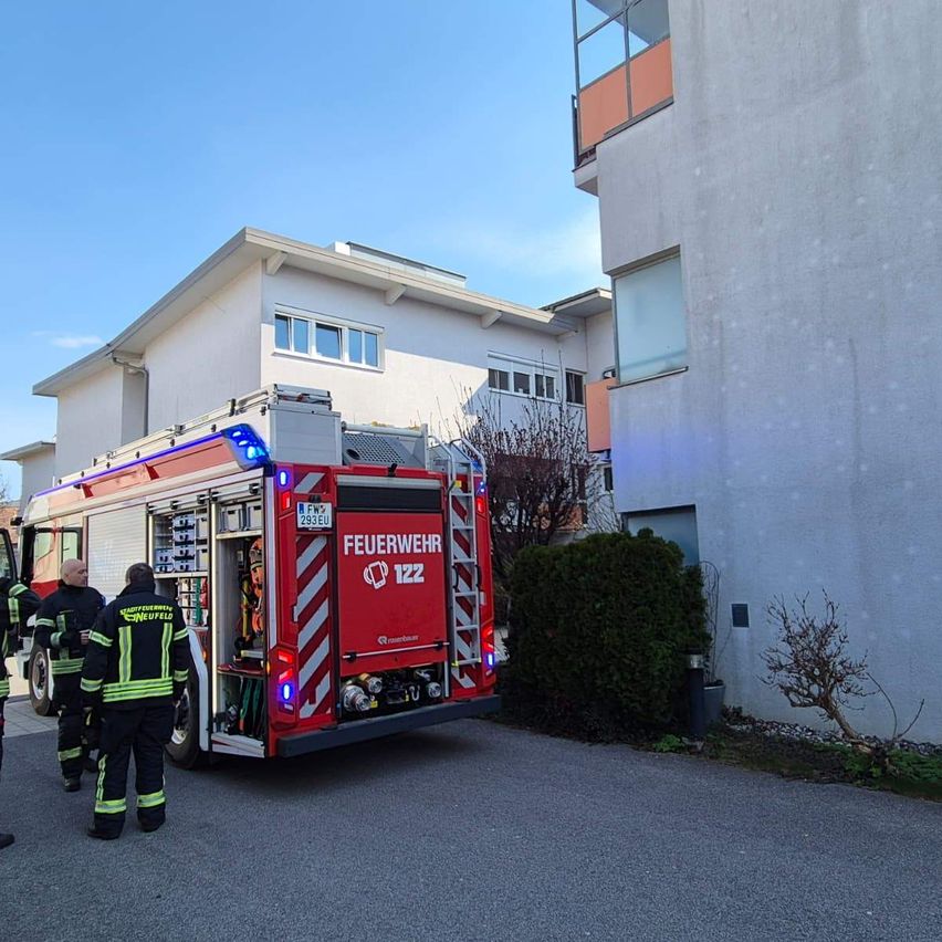 Ein roter Feuerwehrwagen mit eingeschalteten Lichtern ist vor einem Gebäude geparkt. Feuerwehrleute stehen in der Nähe.