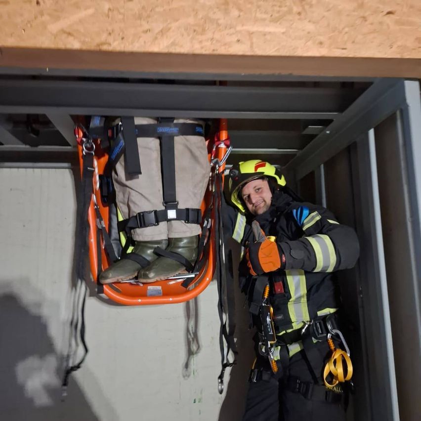 Ein Feuerwehrmann hält eine orange Trage mit einer Person darin, beide sind mit Sicherheitsgurten befestigt. Der Feuerwehrmann trägt einen gelben Helm und einen schwarzen und gelben Sicherheitsanzug.