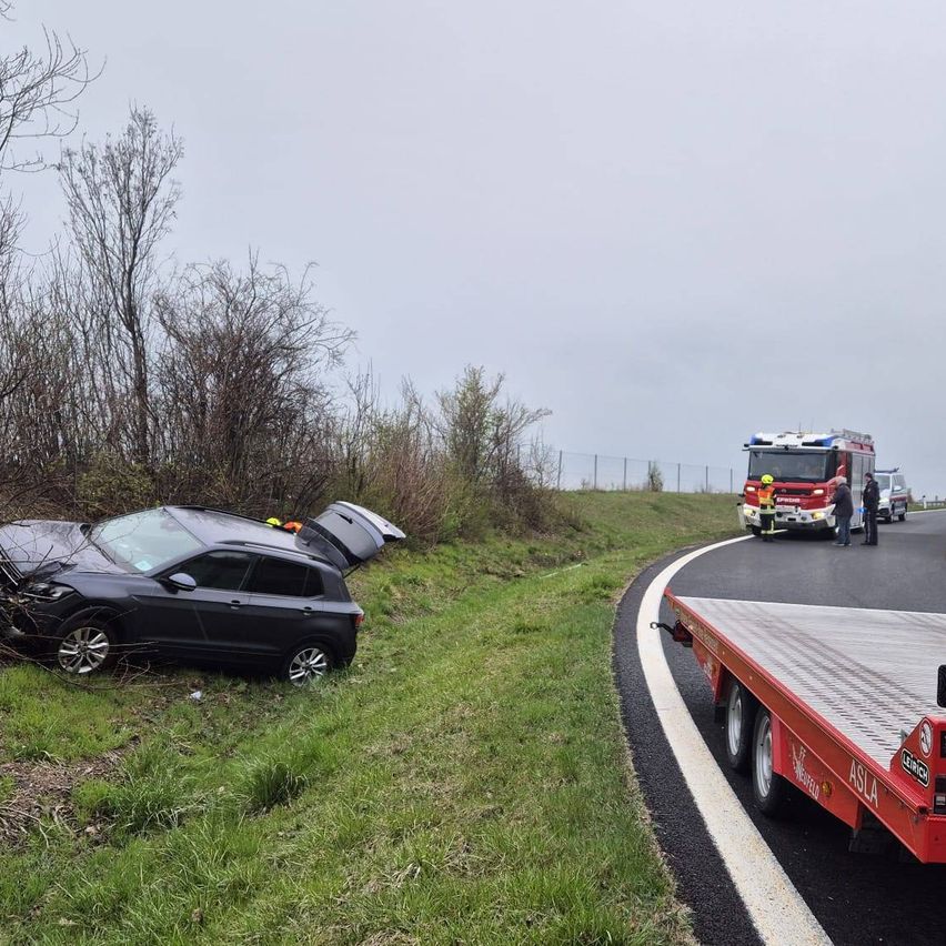 Ein schwarzer SUV ist am Straßenrand in einem Grasfeld zerstört. Zwei Feuerwehrleute stehen neben einem Feuerwehrauto auf der Straße.