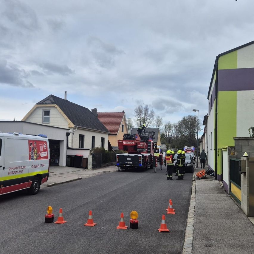 Ein Feuerwehrauto mit Feuerwehrleuten steht auf der Straße. Verkehrskegel markieren das Gebiet. Ein weißer Lieferwagen ist am Straßenrand geparkt.