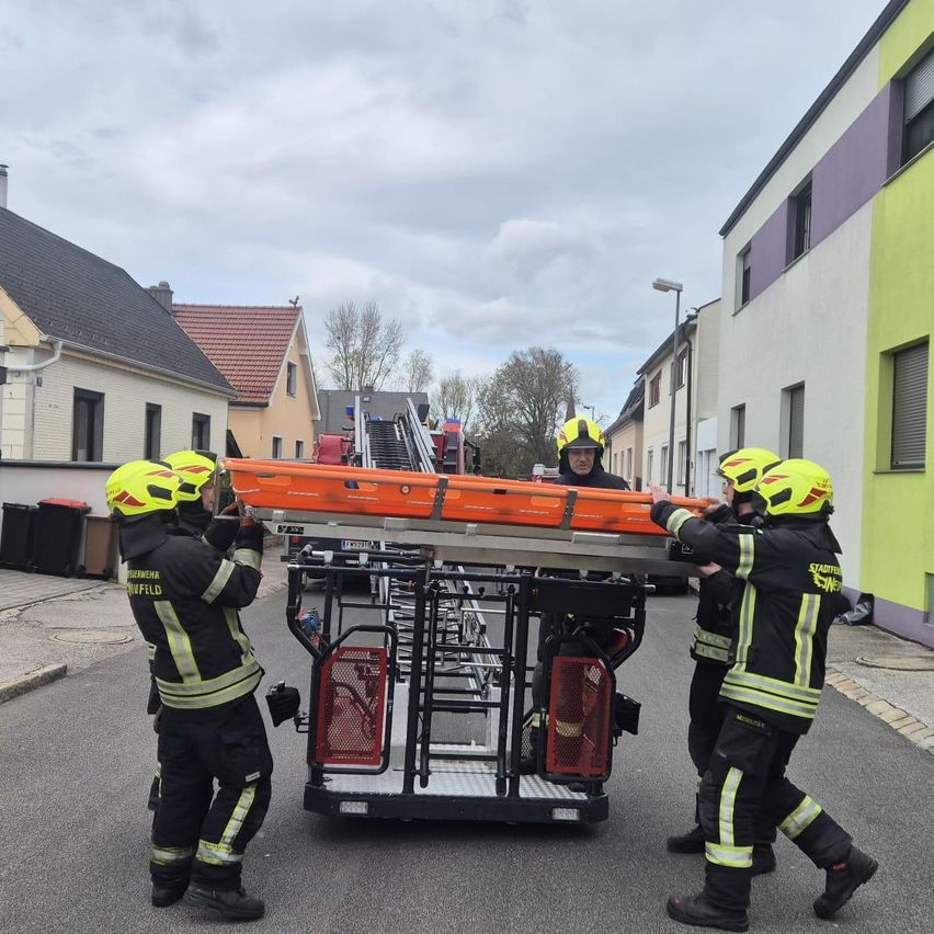 Feuerwehrleute transportieren eine orange Trage auf einer Leiter in einer Straße. Sie tragen Helme und Uniformen. Die Leiter steht auf einem Fahrzeug. Hinter ihnen sind Gebäude mit einigen Mülleimern.