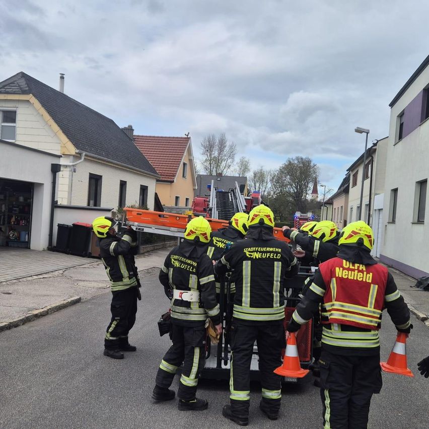 Feuerwehrleute in Schutzkleidung und Helmen stehen um ein Fahrzeug herum. Sie bereiten sich auf einen Notfalleinsatz vor. Die Szene spielt sich auf einer Straße mit Gebäuden auf beiden Seiten ab.