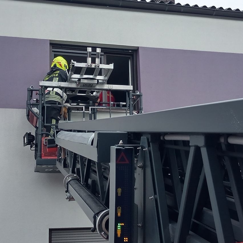 Ein Feuerwehrmann steht auf einer Leiter, trägt einen gelben Helm und Ausrüstung, während eine Person im Fenster eines Gebäudes mit lila und weißen Wänden zu sehen ist.