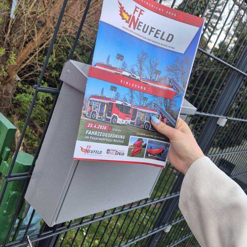 Eine Hand hält ein FF Neufeld-Magazin in der Nähe eines Metallzauns. Das Titelbild des Magazins zeigt einen roten Feuerwehrwagen mit den Texten 'Einladung' und 'Fahrzeugsgung'.