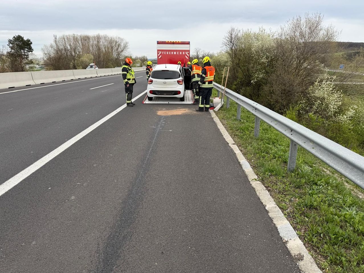 Feuerwehrleute helfen einem weißen Auto am Straßenrand, ein roter Feuerwehrwagen steht in der Nähe.