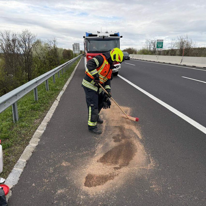 Ein Feuerwehrmann reinigt mit einem Besen eine Verschüttung auf der Straße. Im Hintergrund steht ein Feuerwehrwagen.