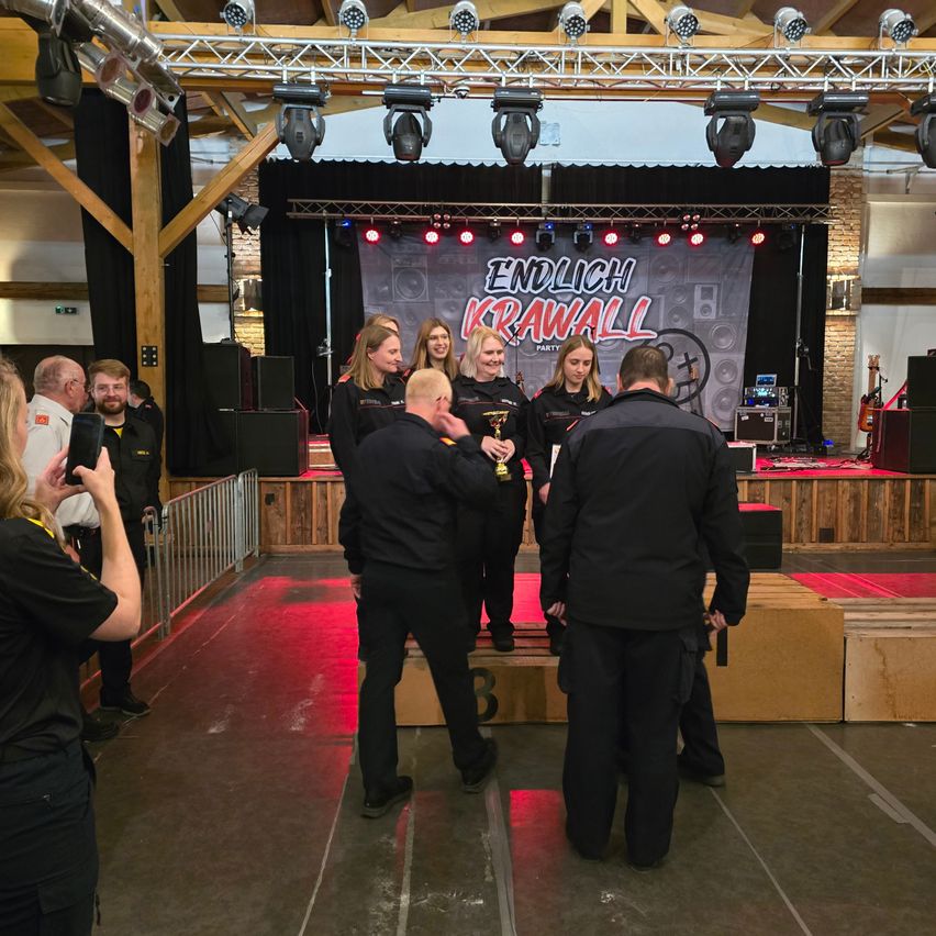 Eine Gruppe von Menschen steht auf einer Bühne mit einem Banner, das 'Endlich Krawall' steht. Sie sind in Schwarz gekleidet, und eine Person hält eine Trophäe. Scheinwerfer sind über ihnen.