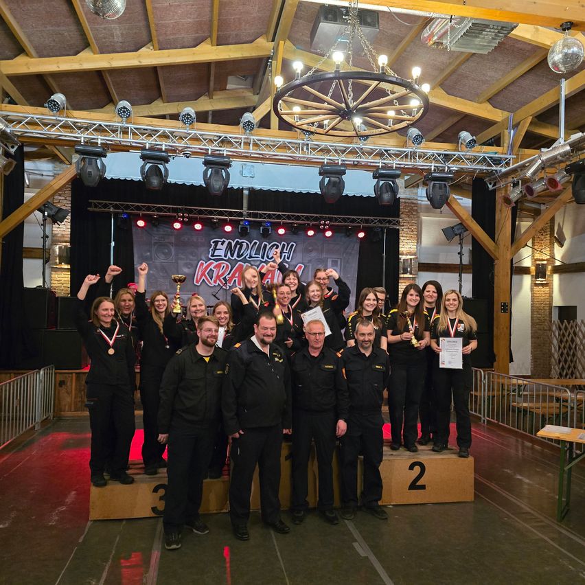 Eine Gruppe von Menschen in gleichen Uniformen steht auf einer Bühne und posiert für ein Foto. Sie halten Medaillen und Urkunden. Die Bühne hat einen Hintergrund mit 'ENDLICH KRALL' darauf. Oben sind ein Kronleuchter und Scheinwerfer zu sehen.