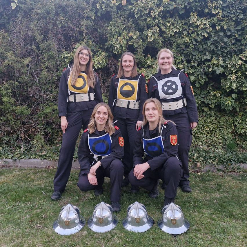 Fünf Frauen in Uniformen mit Helmen posieren vor einem Busch. Sie lächeln und scheinen für ein Foto zu posieren.