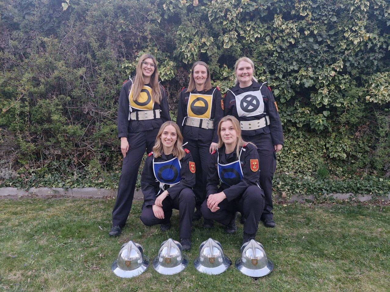 Fünf Frauen in Uniformen mit Helmen posieren vor einem Busch. Sie lächeln und scheinen für ein Foto zu posieren.