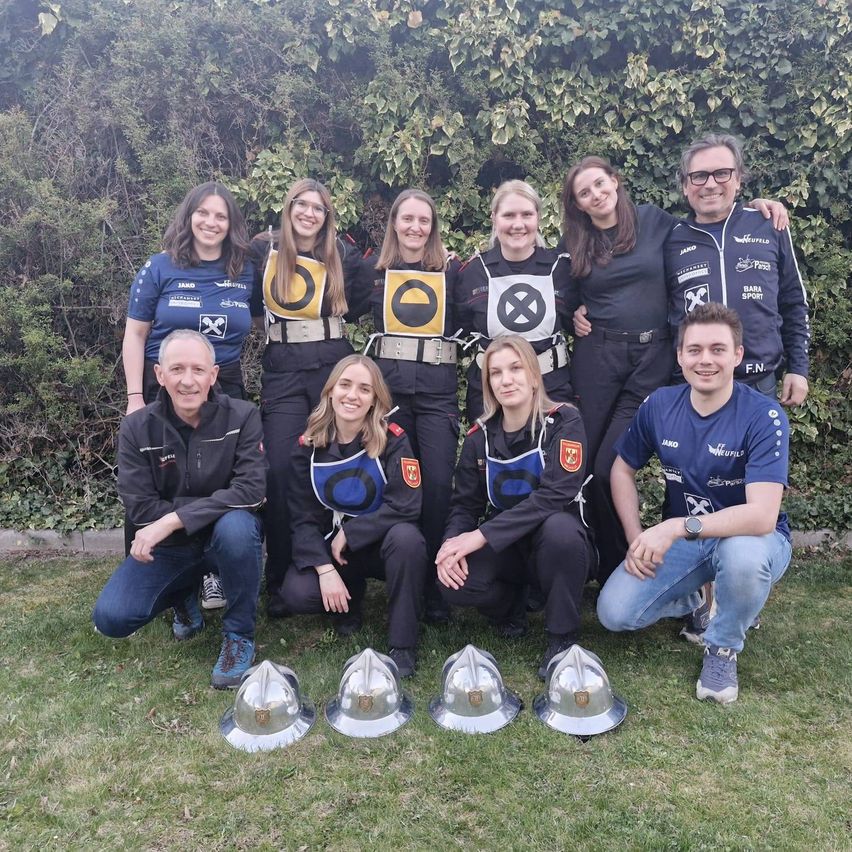 Eine Gruppe von Menschen in Uniform mit Helmen, die lächelnd für ein Foto auf Gras posieren, mit Büschen und Pflanzen im Hintergrund.