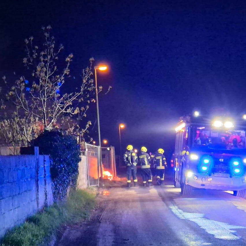 Feuerwehrleute reagieren auf einen Brand in einem ländlichen Gebiet bei Nacht. Das Feuer brennt in der Nähe eines Maschendrahtzauns.