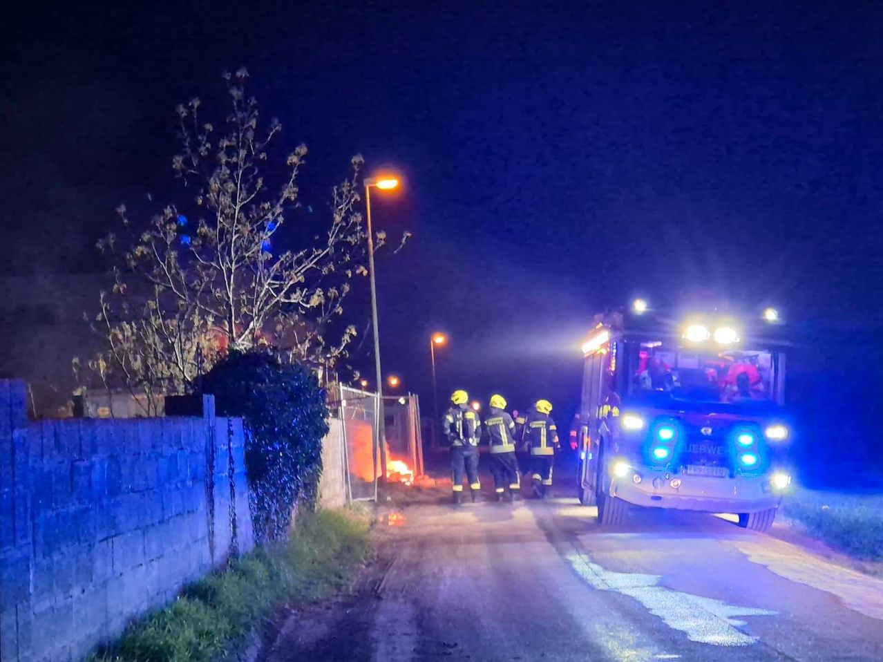 Feuerwehrleute reagieren auf einen Brand in einem ländlichen Gebiet bei Nacht. Das Feuer brennt in der Nähe eines Maschendrahtzauns.
