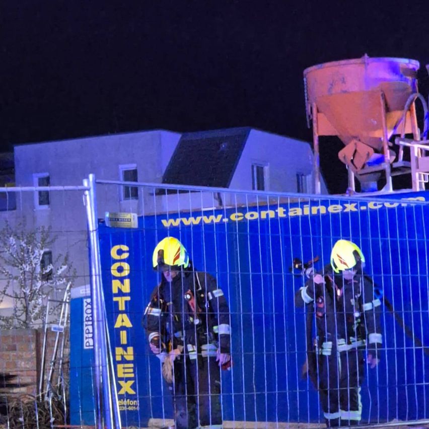 Zwei Feuerwehrleute in voller Ausrüstung stehen hinter einer blauen Barriere mit Containerex.com darauf. Dahinter steht ein hoher Betonmischer. Ein Haus ist im Hintergrund sichtbar.