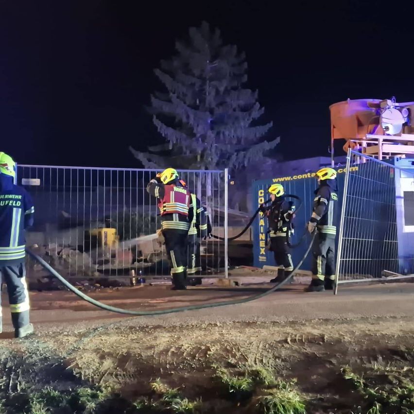 Feuerwehrleute stehen bei einem Metallzaun und halten einen Schlauch. Dahinter ist eine beschädigte Struktur mit einem großen blauen Container zu sehen. Bäume und ein Gebäude sind im Hintergrund.