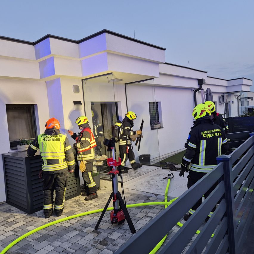 Feuerwehrleute in Schutzkleidung arbeiten vor einem modernen weißen Haus. Sie sind dabei zu sehen, wie sie eine Glastür mit einem Hammer einschlagen. Ein gelber Schlauch ist auf dem Boden zu sehen.