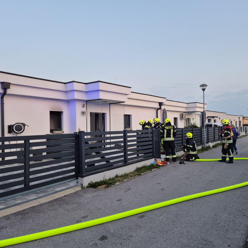 Feuerwehrleute sind vor einem Gebäude mit einem Zaun und löschen einen Brand mit Schläuchen. Ein Feuerwehrmann kniet, während andere in der Nähe stehen.