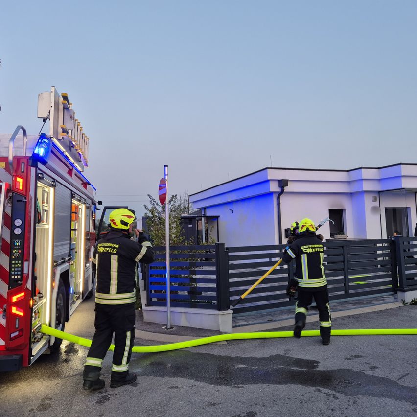 Zwei Feuerwehrleute in voller Ausrüstung, einer mit einer Feuerwehrschläuche, stehen vor einem modernen Gebäude. Ein Feuerwehrwagen mit blinkenden blauen Lichtern ist in der Nähe geparkt.