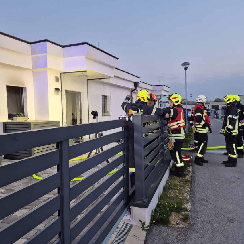 Feuerwehrleute stehen neben einem Tor und halten sich an einem Metallzaun fest, einer öffnet das Tor. Sie stehen vor einem weißen Gebäude mit Glastüren und Fenstern.