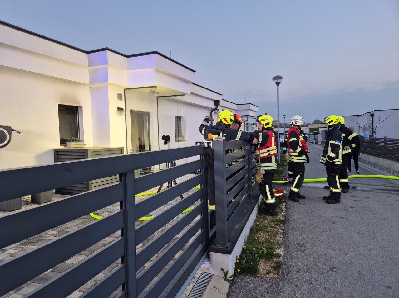 Feuerwehrleute stehen neben einem Tor und halten sich an einem Metallzaun fest, einer öffnet das Tor. Sie stehen vor einem weißen Gebäude mit Glastüren und Fenstern.