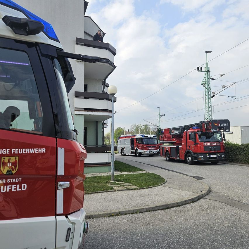 Feuerwehrfahrzeuge sind neben einem Gebäude mit Balkon geparkt. Eine Person steht auf dem Balkon und eine Straßenlaterne ist in der Nähe. Die Straße hat Gras am Rand.