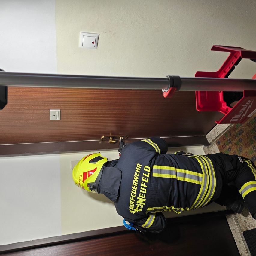 Ein Feuerwehrmann in einem gelben Helm und blauen Hosen arbeitet mit einem Werkzeug an einer Tür.