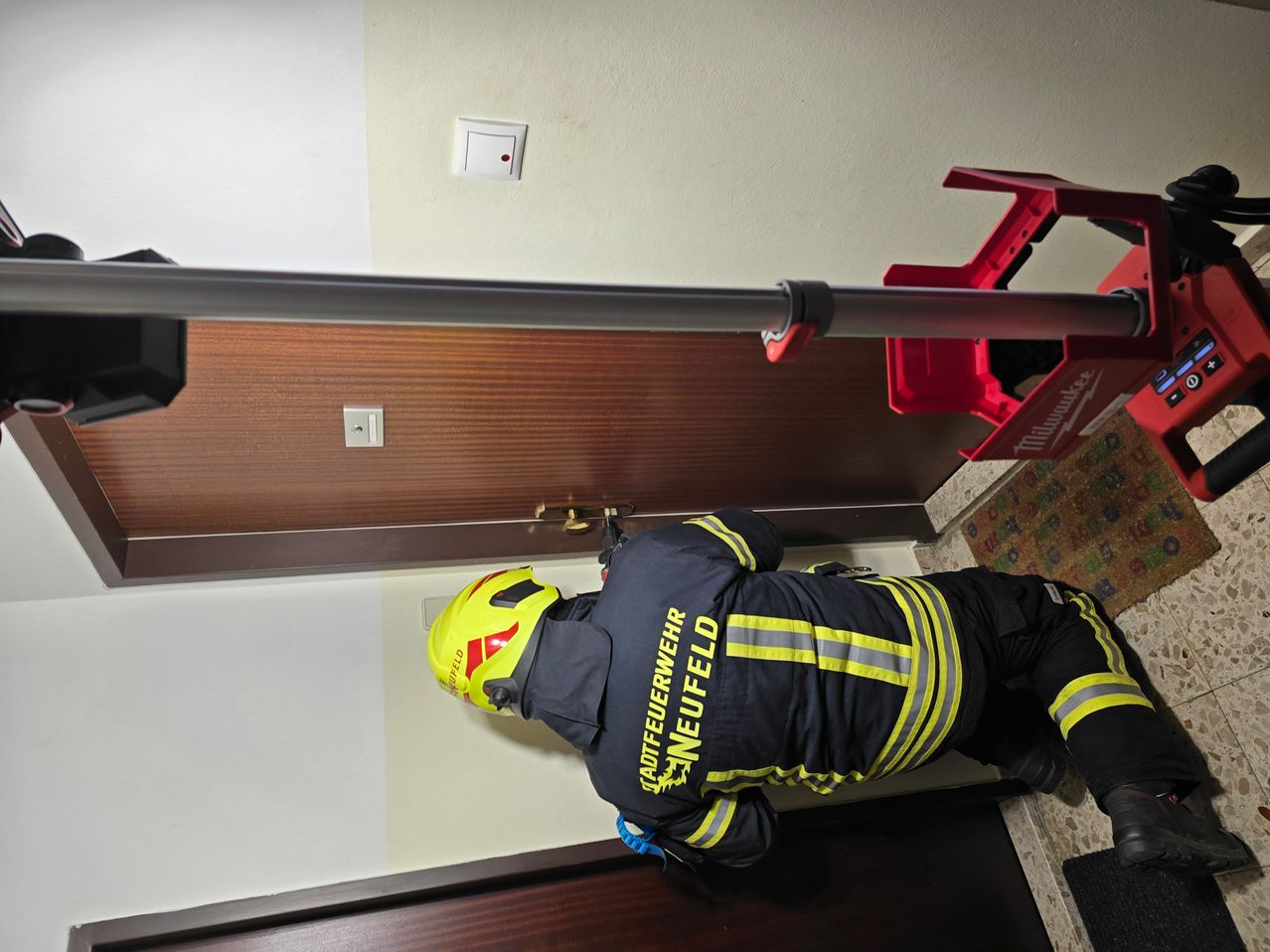 Ein Feuerwehrmann in einem gelben Helm und blauen Hosen arbeitet mit einem Werkzeug an einer Tür.