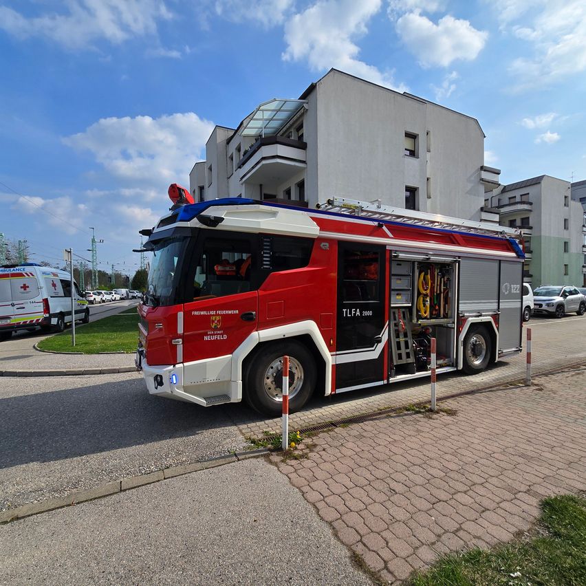 Ein roter Feuerwehrwagen mit Leiter steht auf einem gepflasterten Bereich in der Nähe von Gebäuden unter einem teilweise bewölkten Himmel.