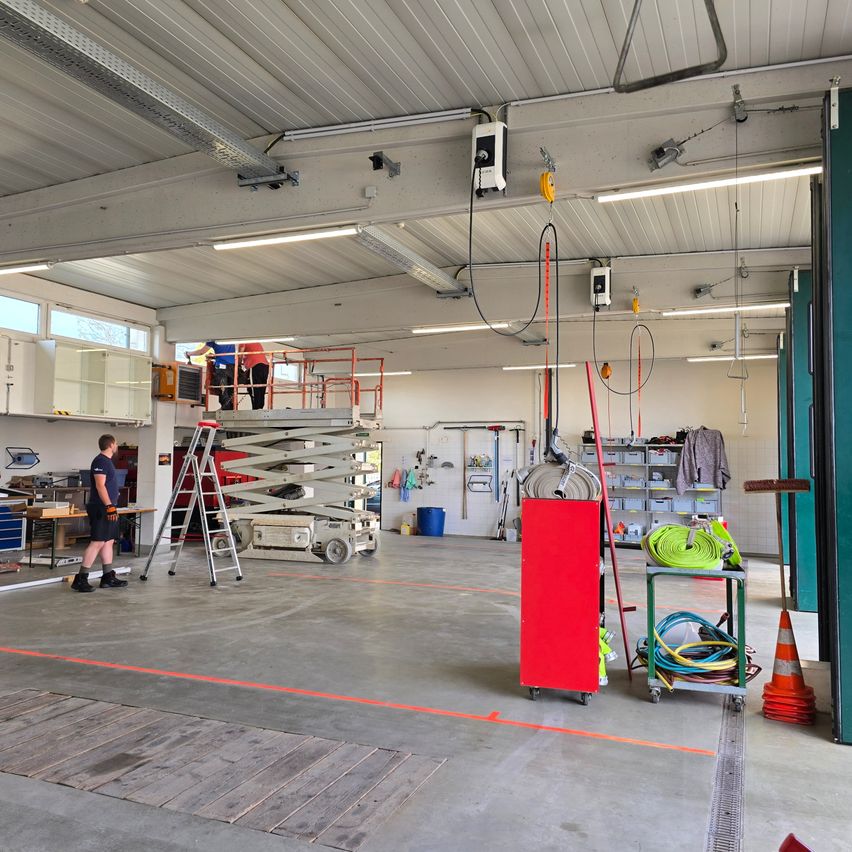 Ein geräumiger Werkstattraum mit Arbeitern auf einem Hebebühne. Ein Mann steht neben einer Leiter. Verschiedene Werkzeuge und Geräte sind auf Regalen und Wagen gelagert. Der Boden hat orange Markierungen.