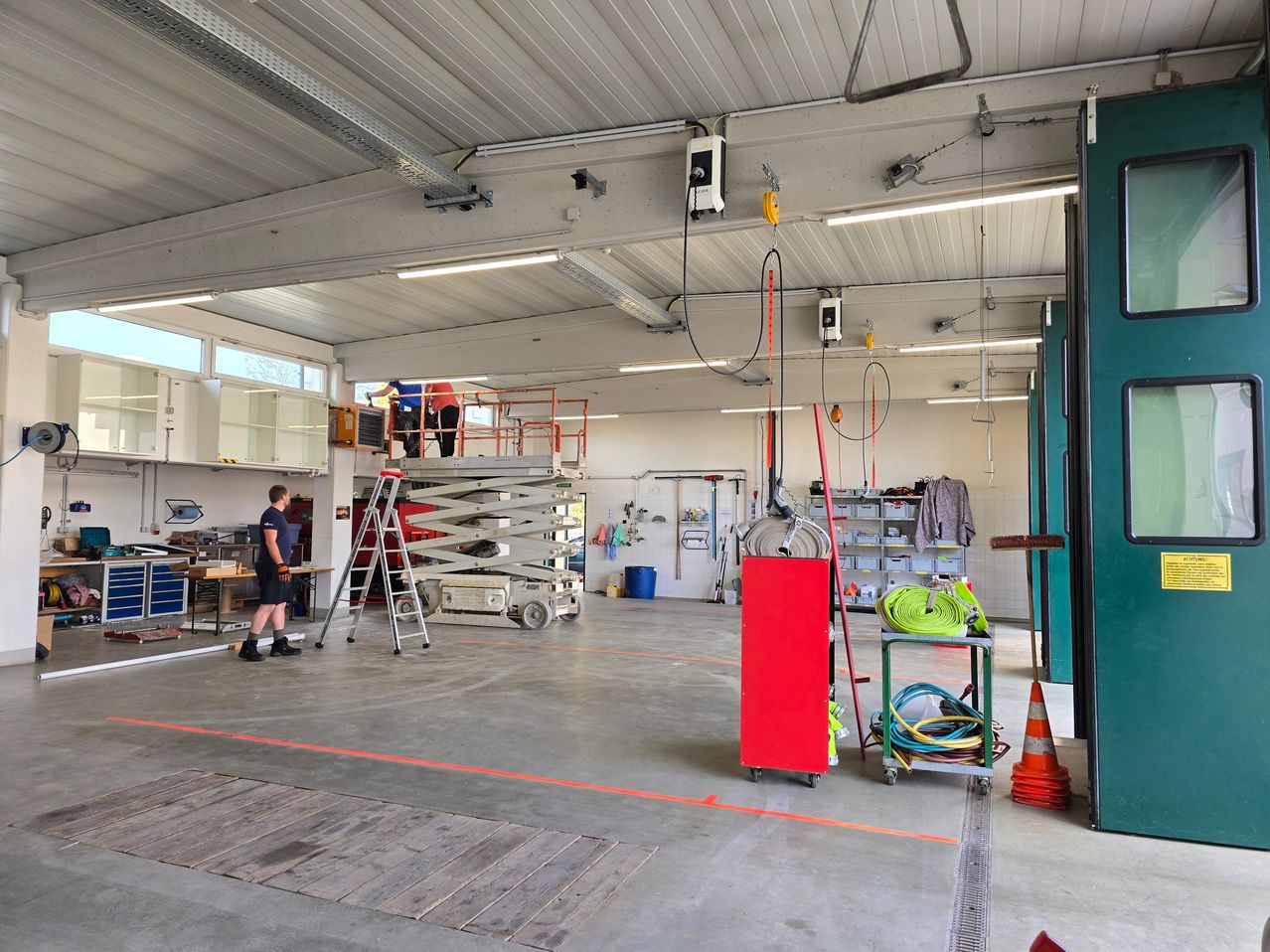Ein geräumiger Werkstattraum mit Arbeitern auf einem Hebebühne. Ein Mann steht neben einer Leiter. Verschiedene Werkzeuge und Geräte sind auf Regalen und Wagen gelagert. Der Boden hat orange Markierungen.