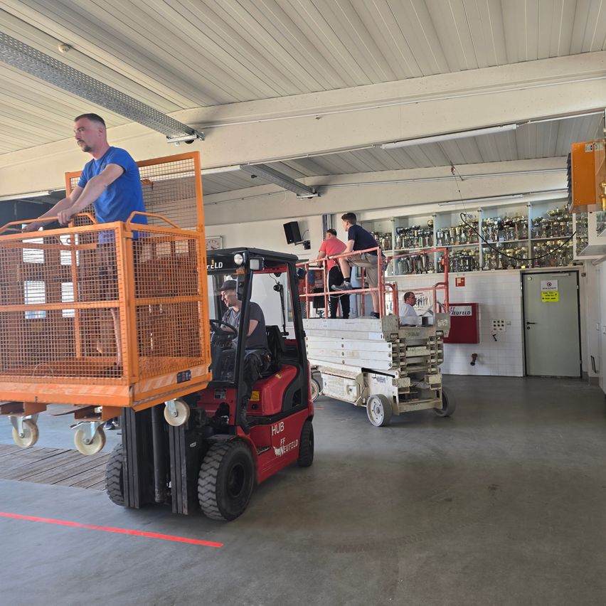 Ein Gabelstapler mit einem Korb wird in einem Lagerhaus gefahren, während Arbeiter auf einer Gerüstarbeit arbeiten. Ein Lagerkasten und andere Geräte sind in der Nähe.