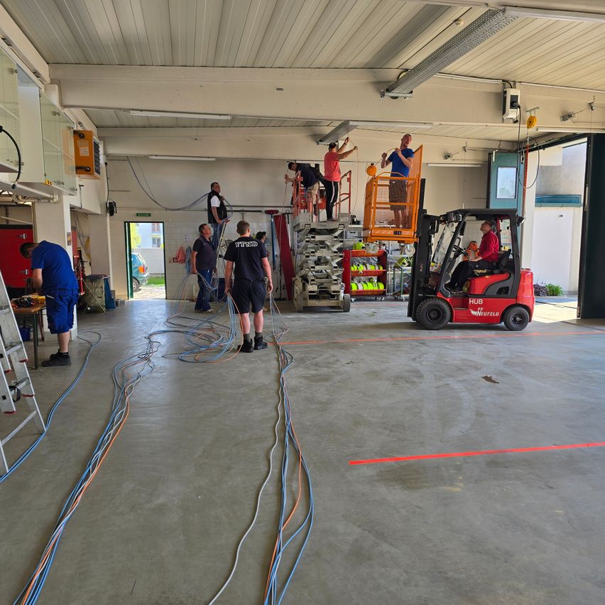 In einer Werkstatt arbeiten Menschen an einem Projekt. Zwei Personen stehen auf einer Leiter, eine fährt einen Gabelstapler und andere sind am Boden. Drähte sind mit der Decke verbunden, und eine Leiter steht in der Nähe.