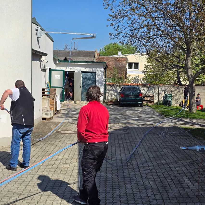 Zwei Männer arbeiten an einem sonnigen Tag vor einem Haus. Ein Mann zieht einen Schlauch, während der andere zusieht. Ein Auto ist auf der Einfahrt geparkt.