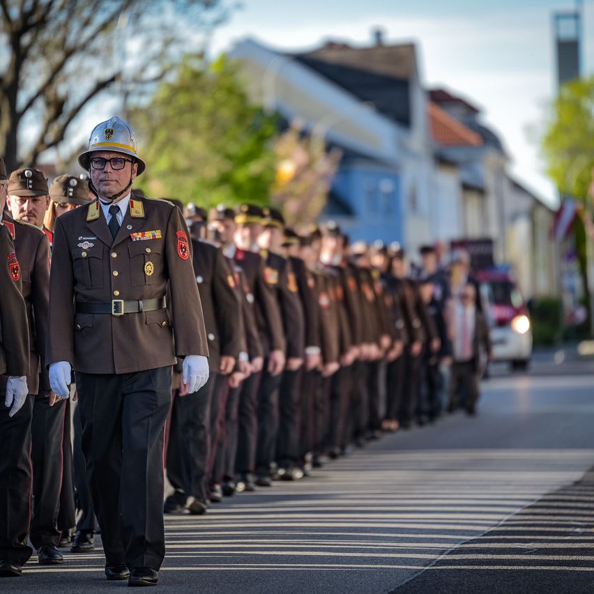 Eine Militärparade bewegt sich eine Straße hinunter, mit Bäumen und Gebäuden im Hintergrund. Uniformierte Soldaten in identischer Kleidung marschieren im Gleichschritt.