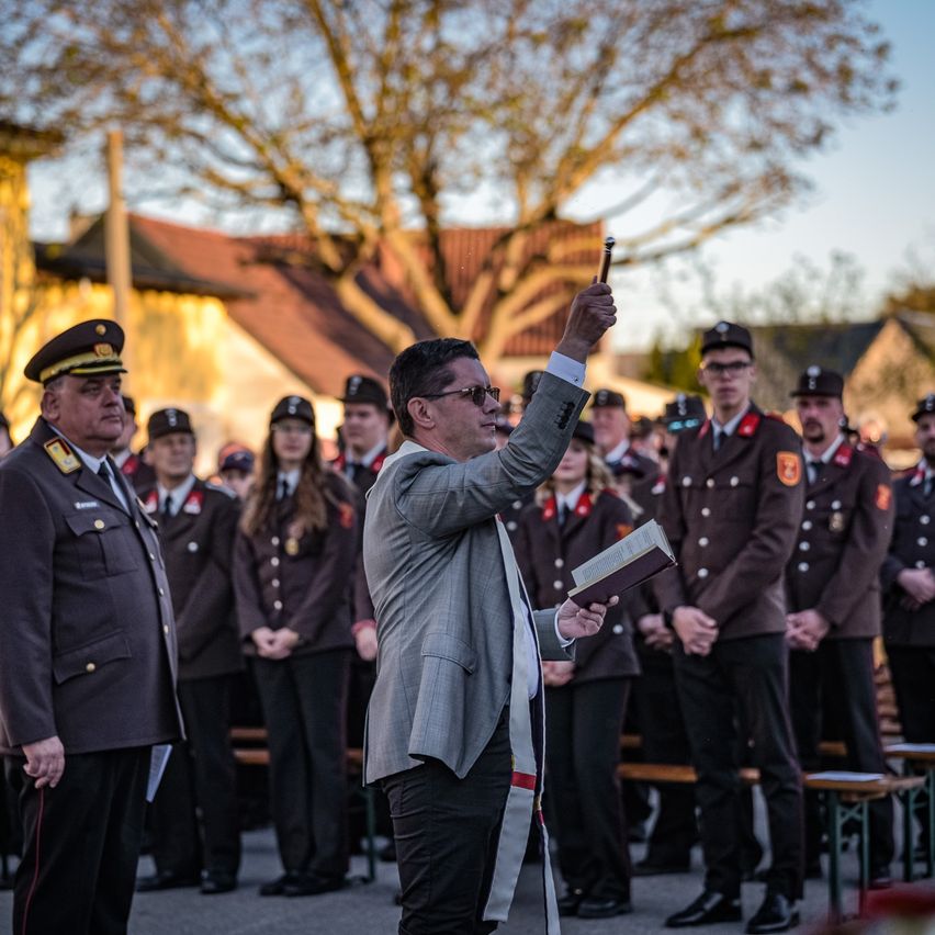 Ein Mann in einem grauen Anzug hält ein Buch und einen Stift und steht vor einer Gruppe uniformierter Personen.